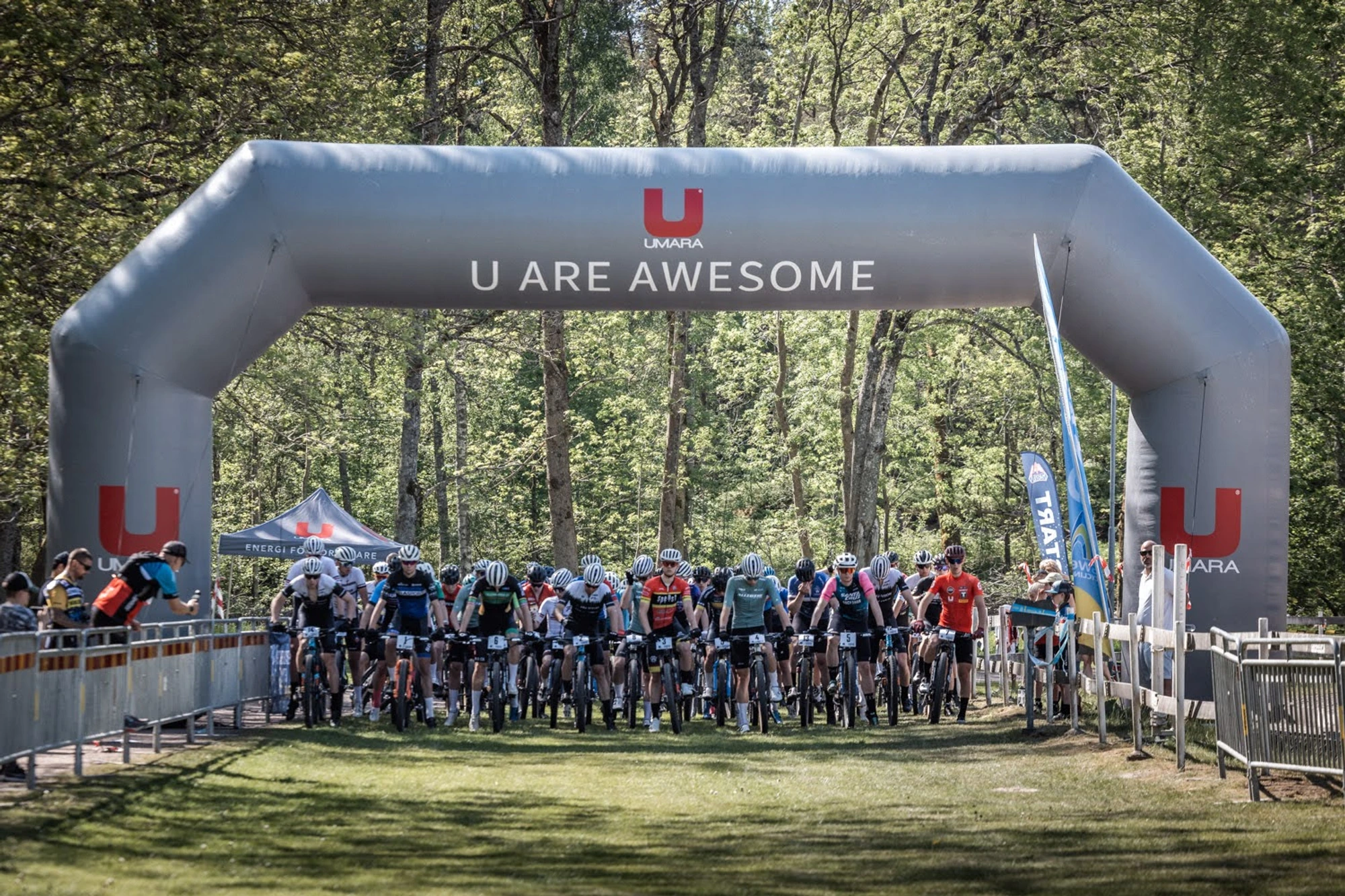 Ett cykellopp där deltagare står redo vid startlinjen under en båge som bär texten "U ARE AWESOME". I bakgrunden syns träd.