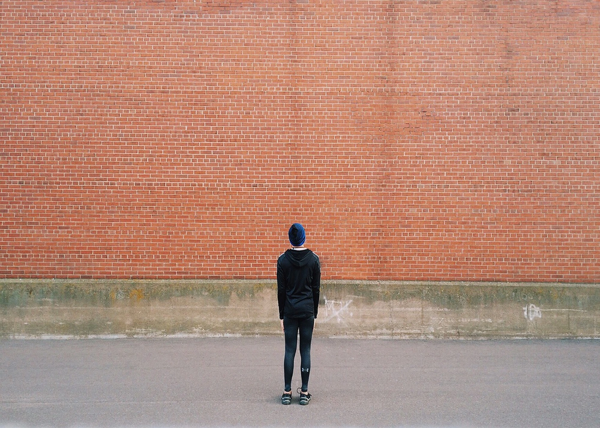 En person står vänd bort från kameran framför en stor, röd tegelvägg, iförd mörka kläder och en mössa.