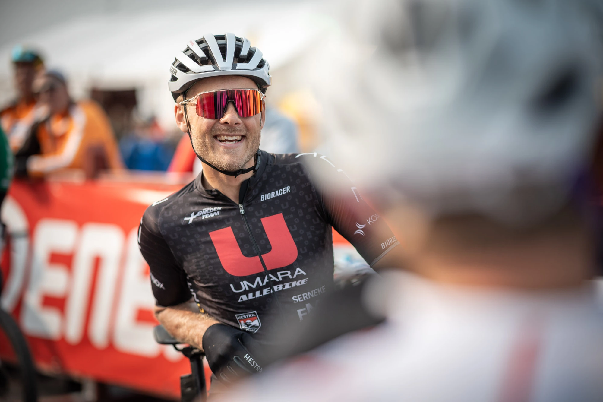 En cyklist klädd i svart, med hjälm på huvudet, ler stort under ett cykellopp. De har solglasögon på sig och står framför en banderoll i röd färg med text på. I bakgrunden kan man urskilja andra människor och cyklister.