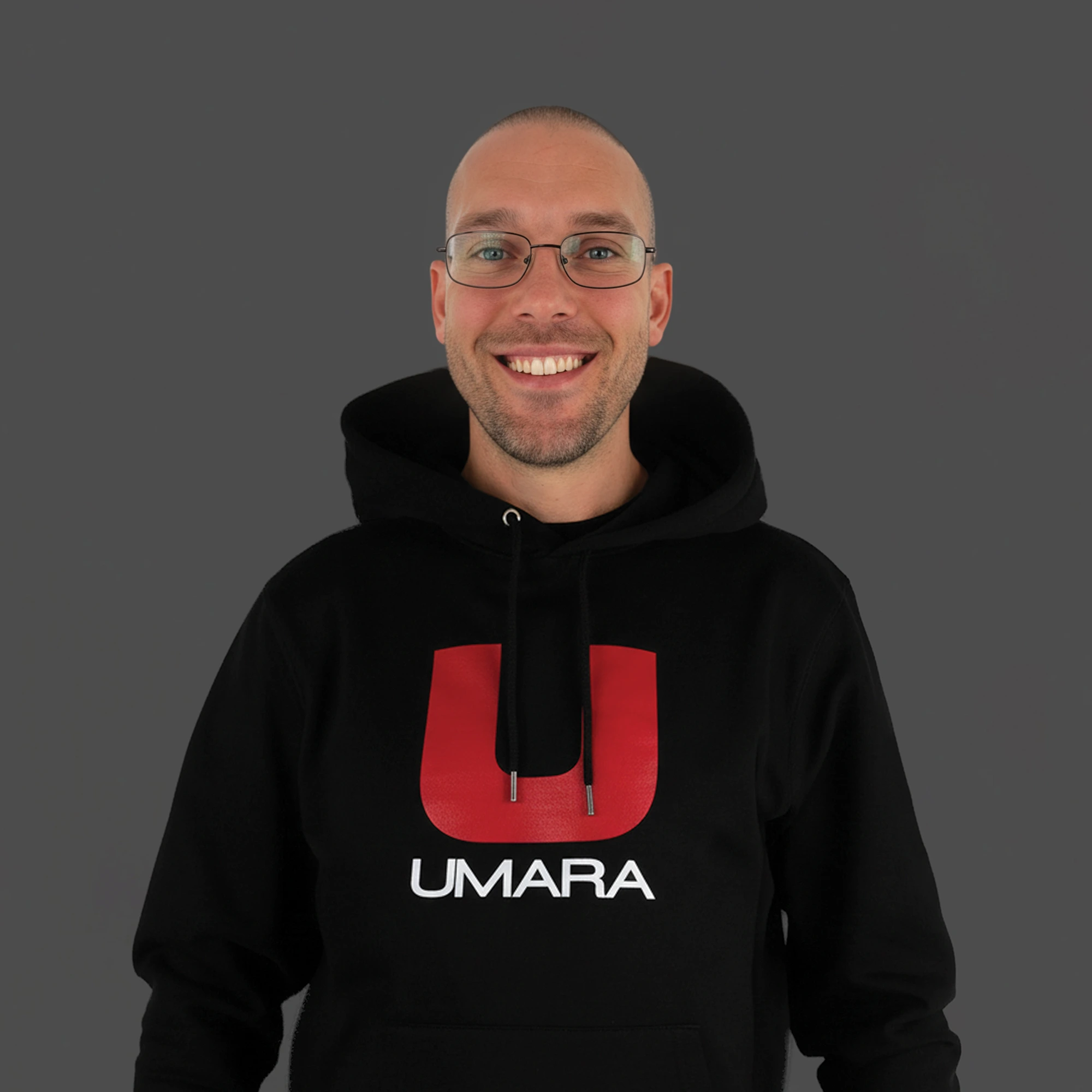 En person med glasögon bär en svart hoodie prydd med texten "UMARA" och ett stort rött "U" på framsidan. Personen står mot en grå bakgrund.