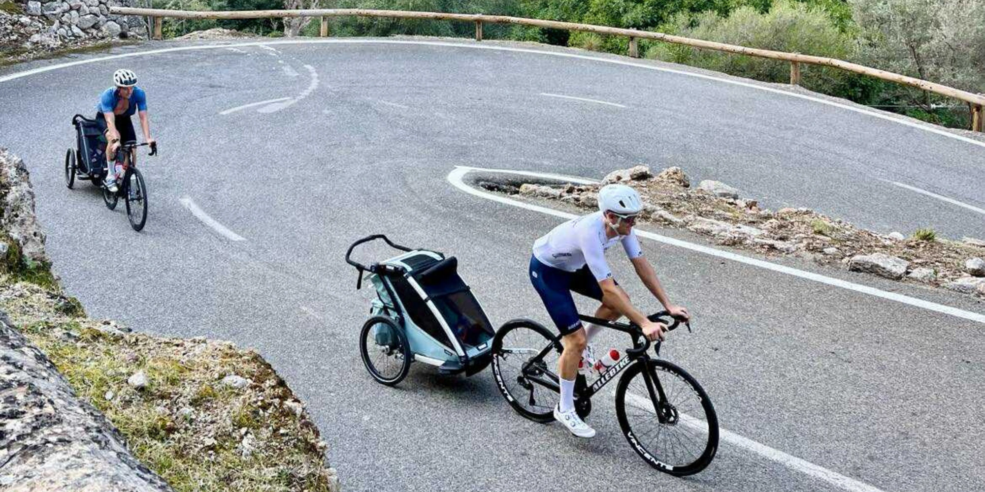 Två cyklister följer en slingrande väg, där den främre drar en barnvagn som är kopplad till sin cykel.