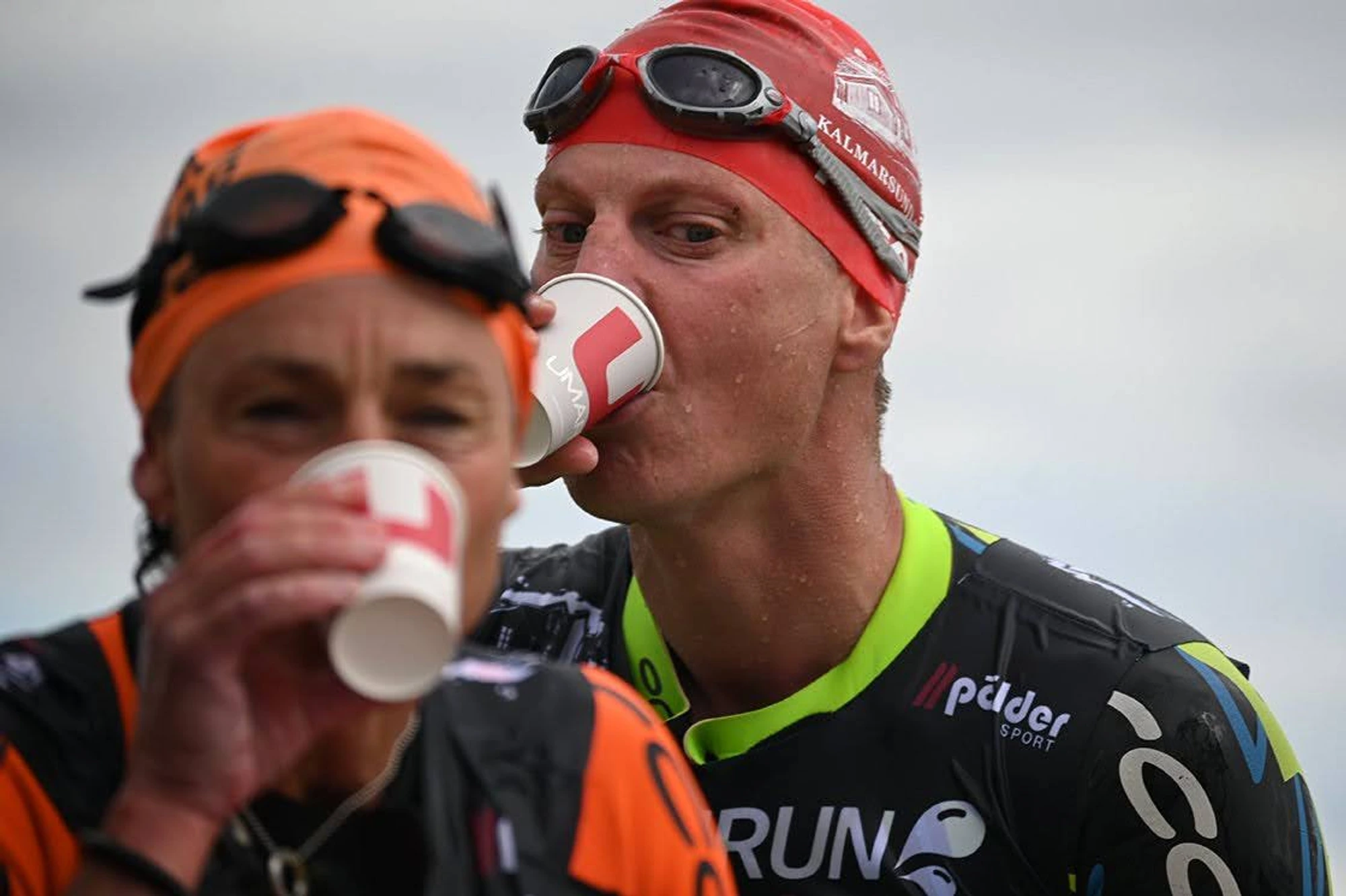 Under en tävling ses två idrottare dricka från pappersmuggar, klädda i simglasögon och våtdräkter.