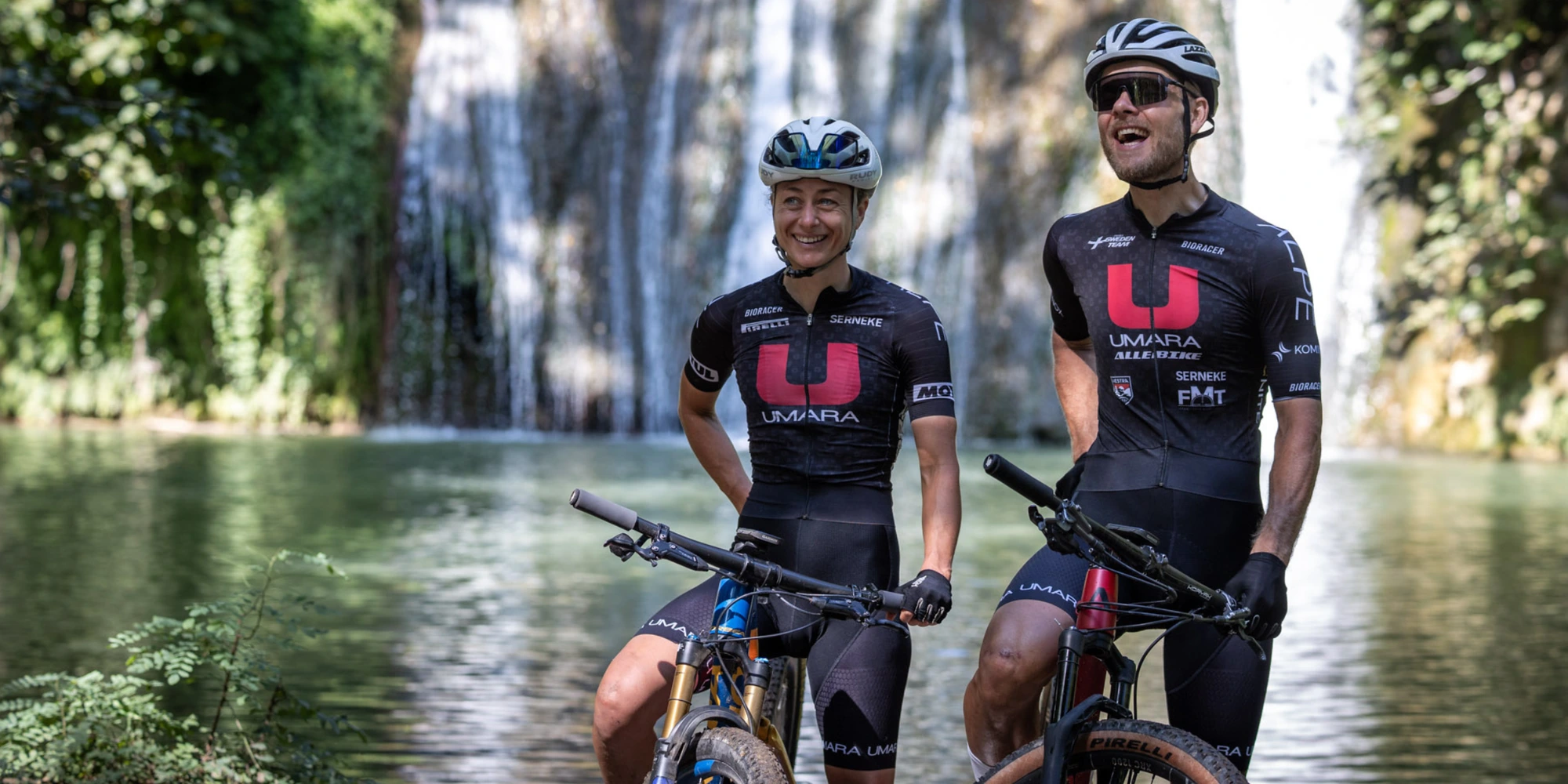 Två cyklister klädda i UCI-uniformer står framför ett vattenfall med sina mountainbikes.