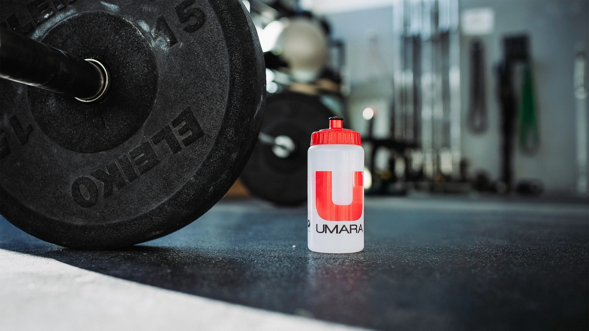 A white water bottle with a red cap, adorned with the "UMARA" logo, sits on a gym floor next to a barbell. Exercise equipment and weights can be glimpsed in the background.